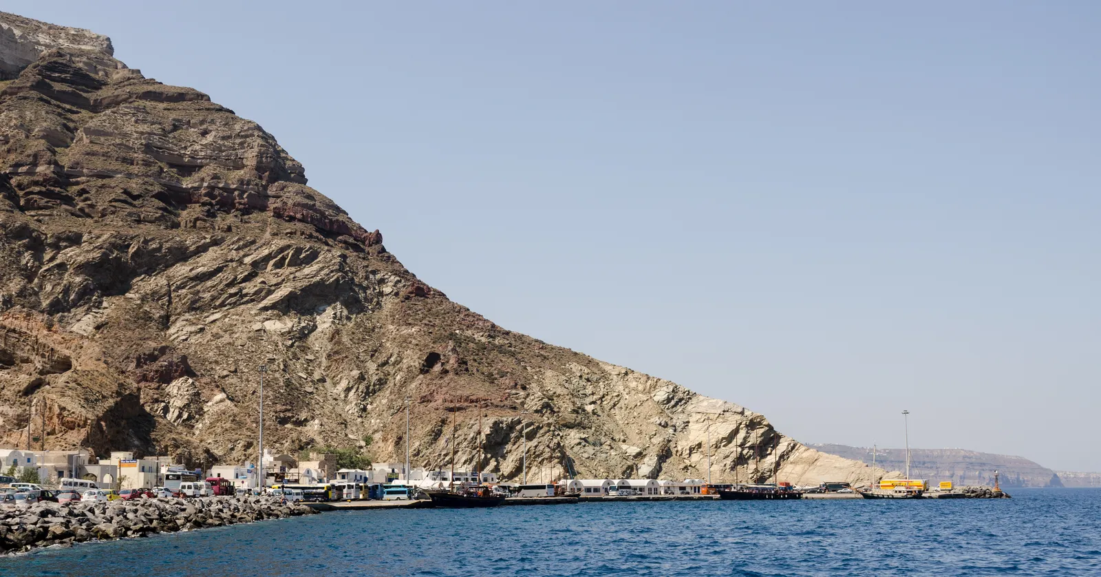 Athinios port from the water, a single strip of quay with parked cars and ferry berths pressed against the base of a steep brown cliff with no town behind it