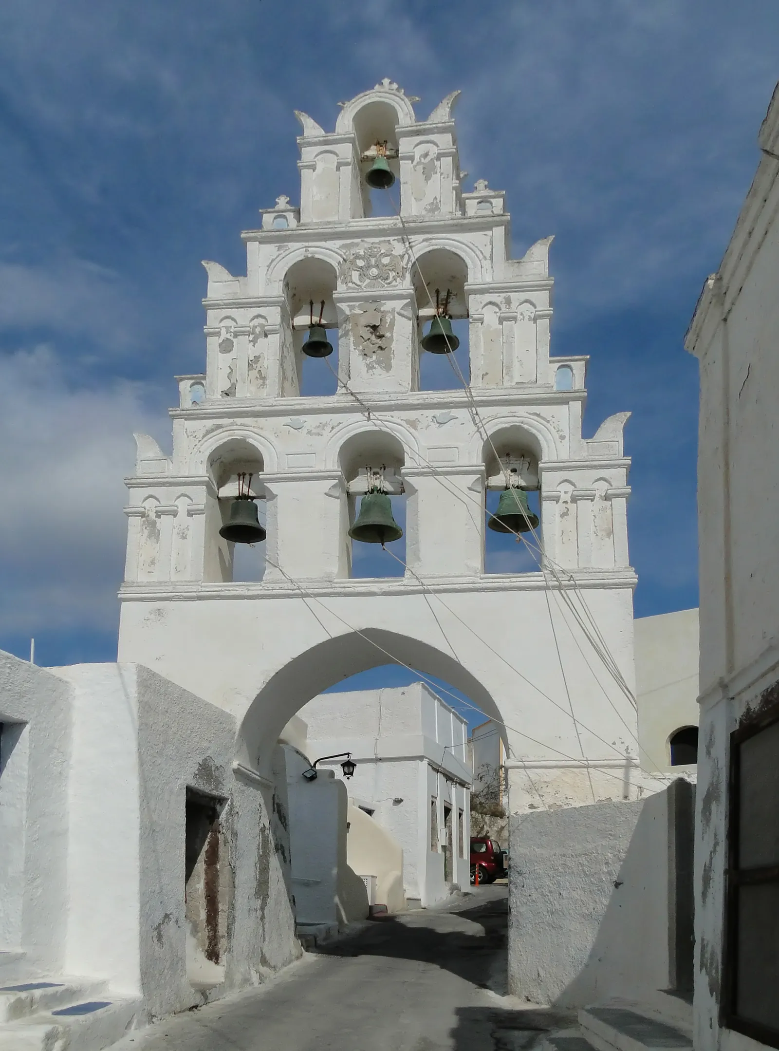 A tall white bell tower in Megalochori built as a tiered wall of arches holding six bells, standing over an archway across a narrow lane