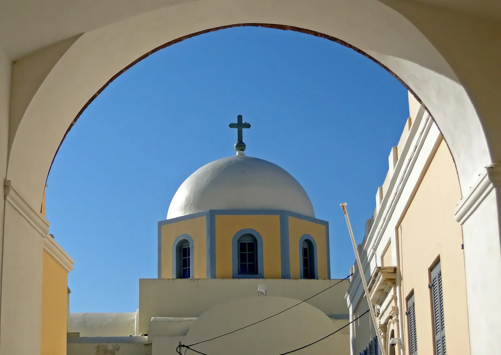 The pale yellow and white dome of Fira's Catholic cathedral framed through a whitewashed street arch, its blue-trimmed windows below the drum