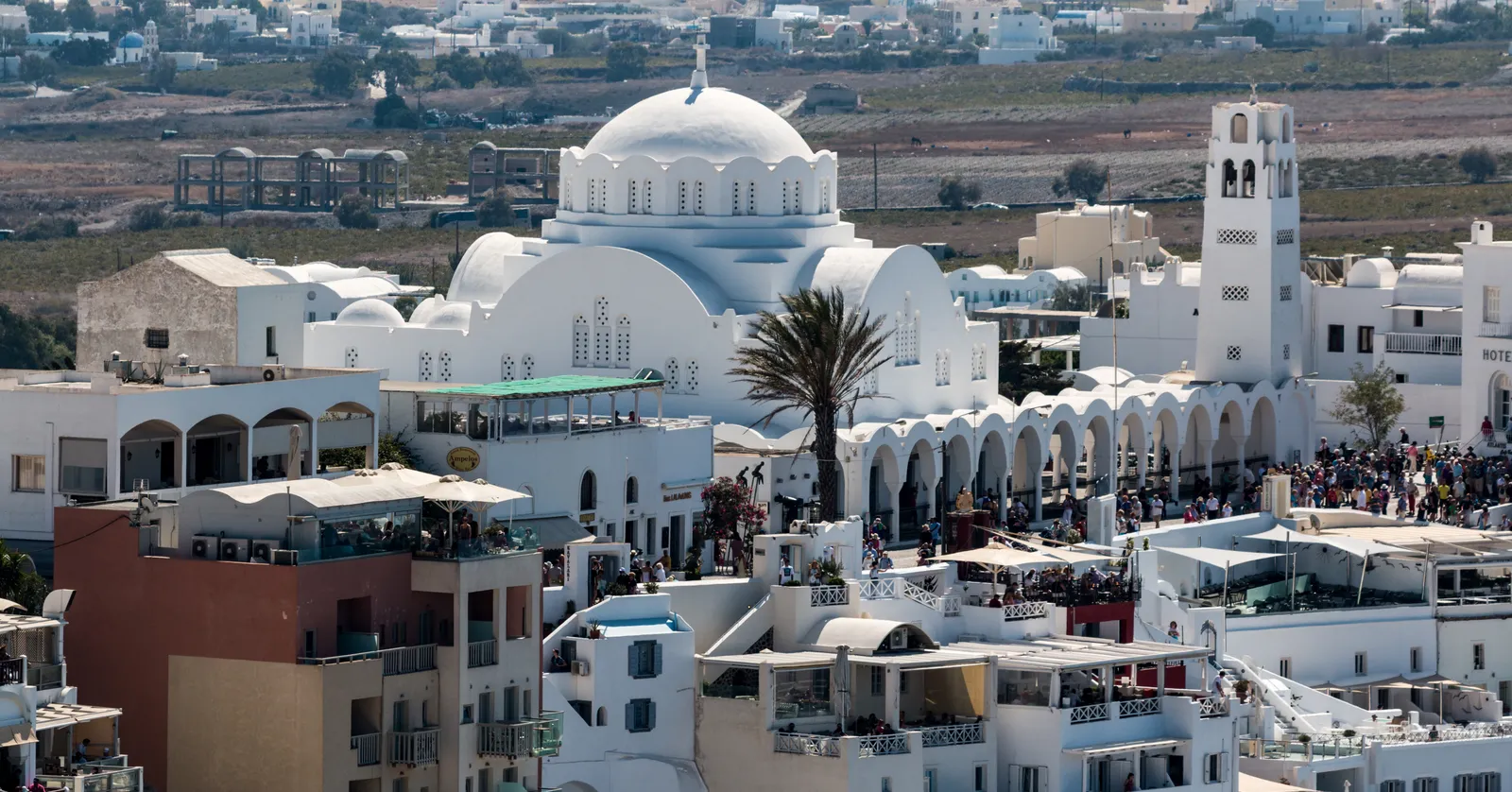 Central Fira from above, a dense white town of terraces and domes with a bell tower on the right and crowds filling the walkways between the cafes