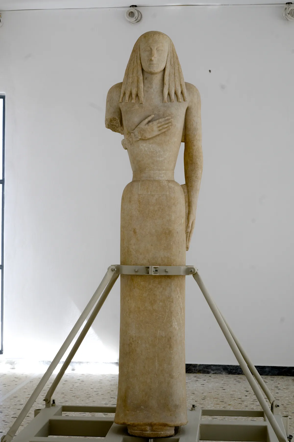 The Kore of Thera, a marble figure of a young woman with long braided hair and one hand at her chest, standing on a metal frame in a plain white museum room