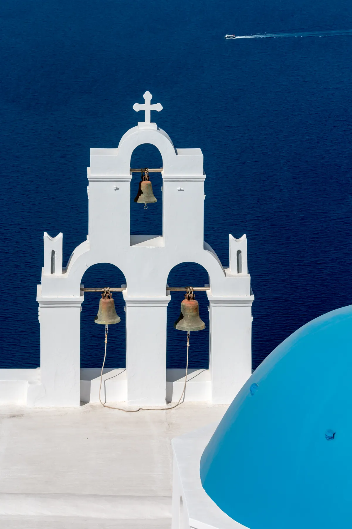 The Three Bells of Fira, a white belfry with three bronze bells hung in stepped arches on a rooftop, with the blue dome of the church beside it and the caldera water far below
