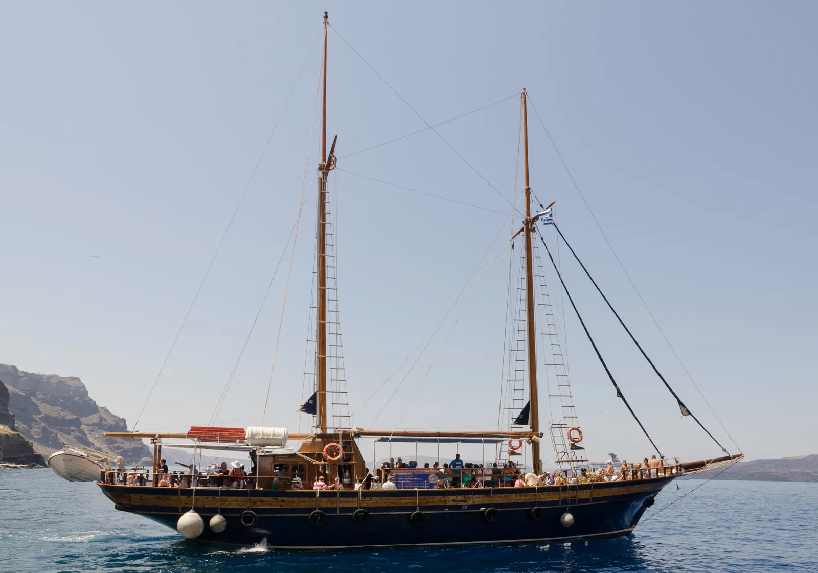 A two-masted wooden excursion boat crowded with passengers on the open deck, motoring across the caldera off Santorini