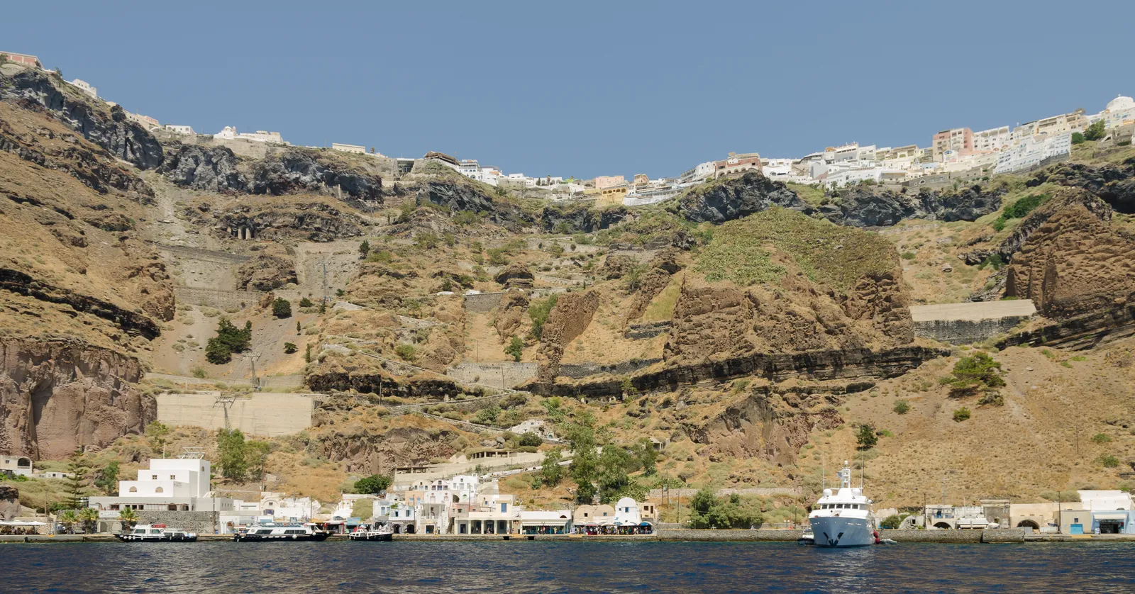 Fira's Old Port seen from the water, a thin line of white quayside buildings at the foot of a banded volcanic cliff with the town on the rim far above and a boat moored alongside