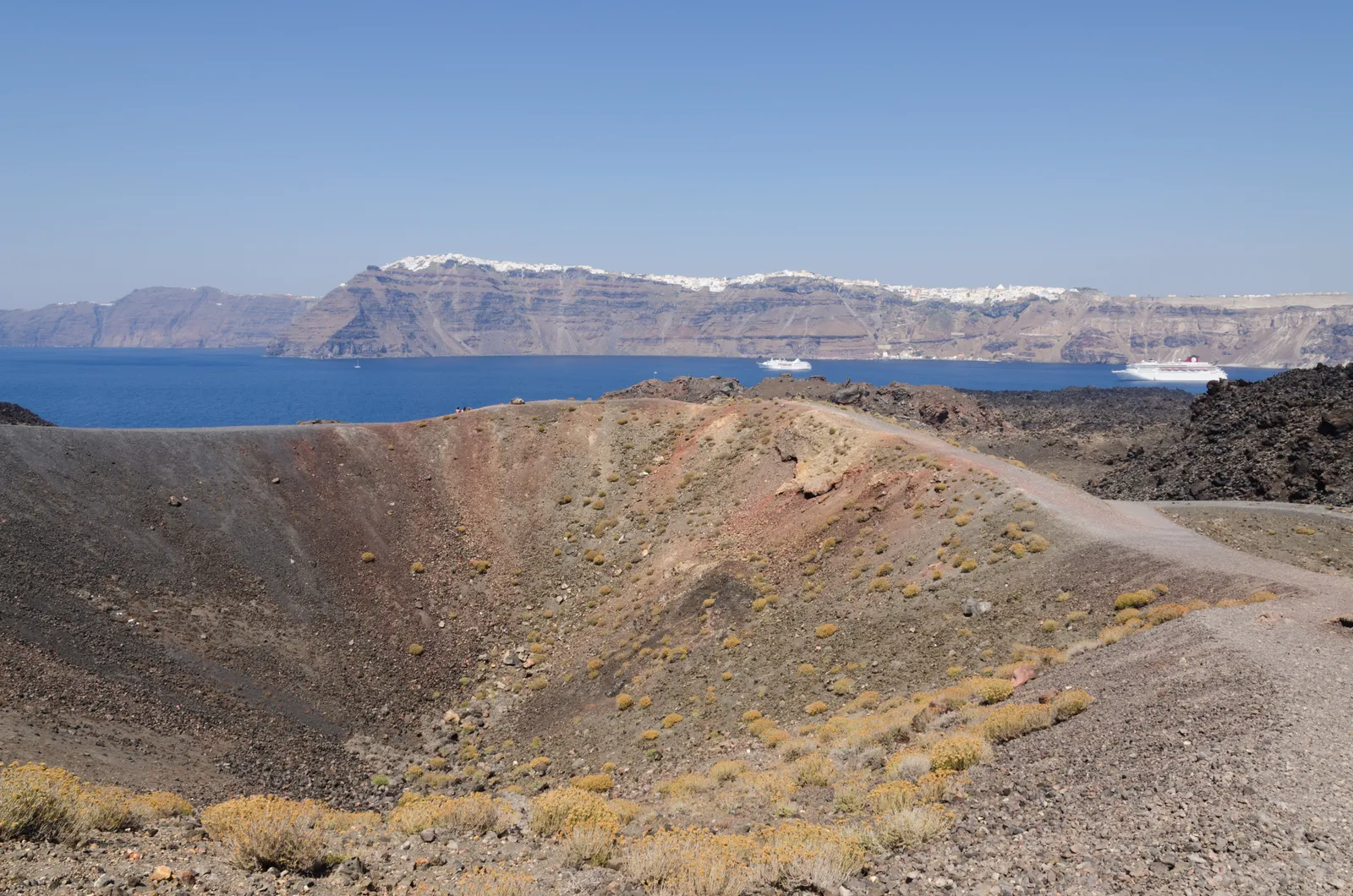 The rust-red crater rim of Nea Kameni with a gravel path running along it, the blue caldera beyond and the white line of Santorini's clifftop villages on the far wall