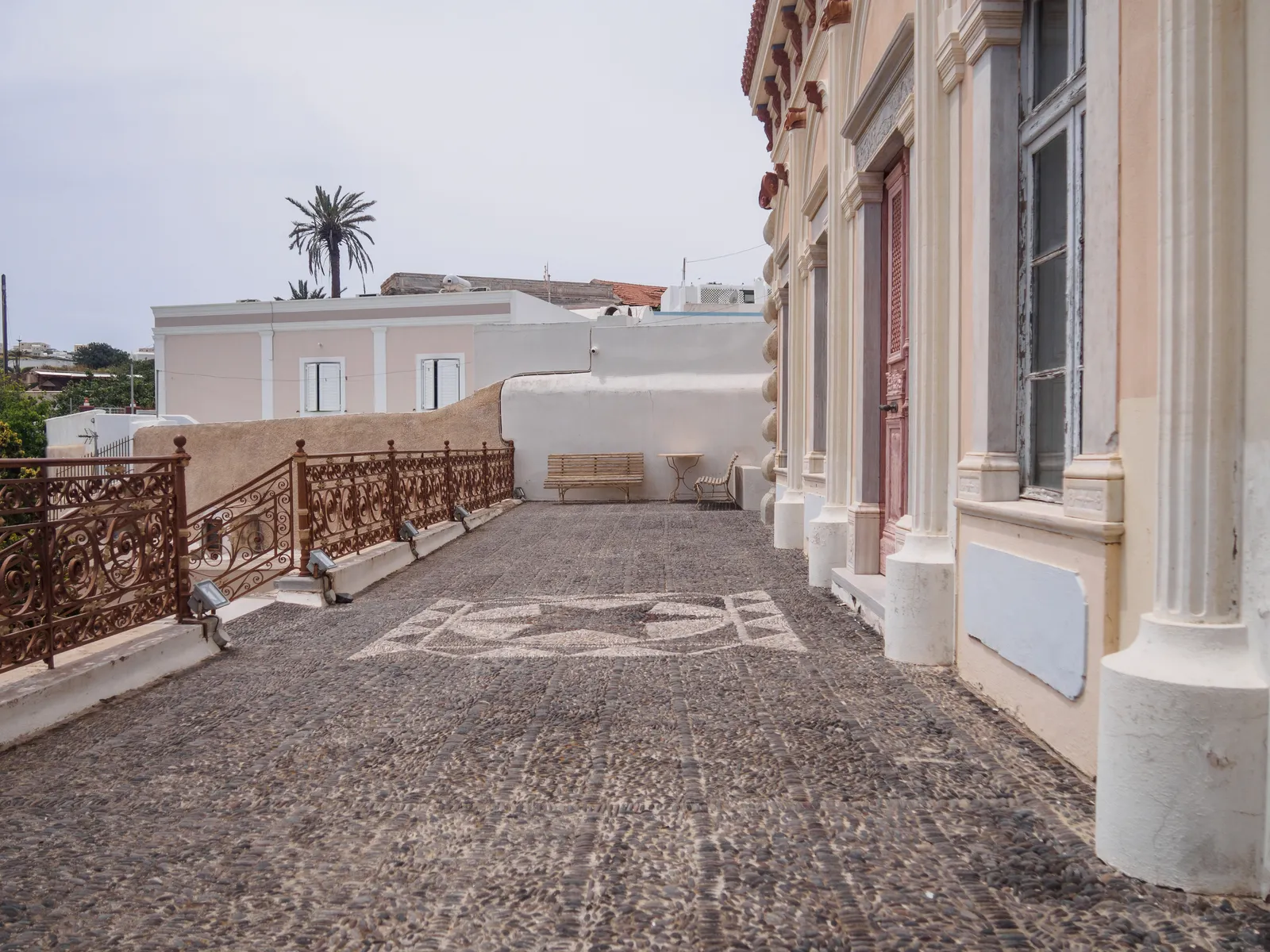 The terrace of the Argyros mansion in Messaria, a patterned pebble mosaic floor running beside a pastel neoclassical facade with cast-iron railings along the open side