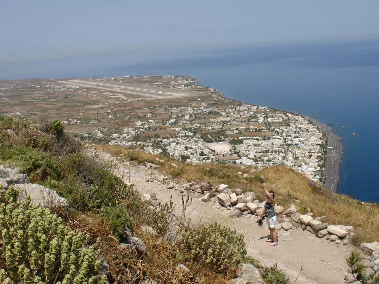Santorini's single runway seen from the ridge above, running parallel to the coast with the flat farmland of the island's east side on one side and the sea on the other