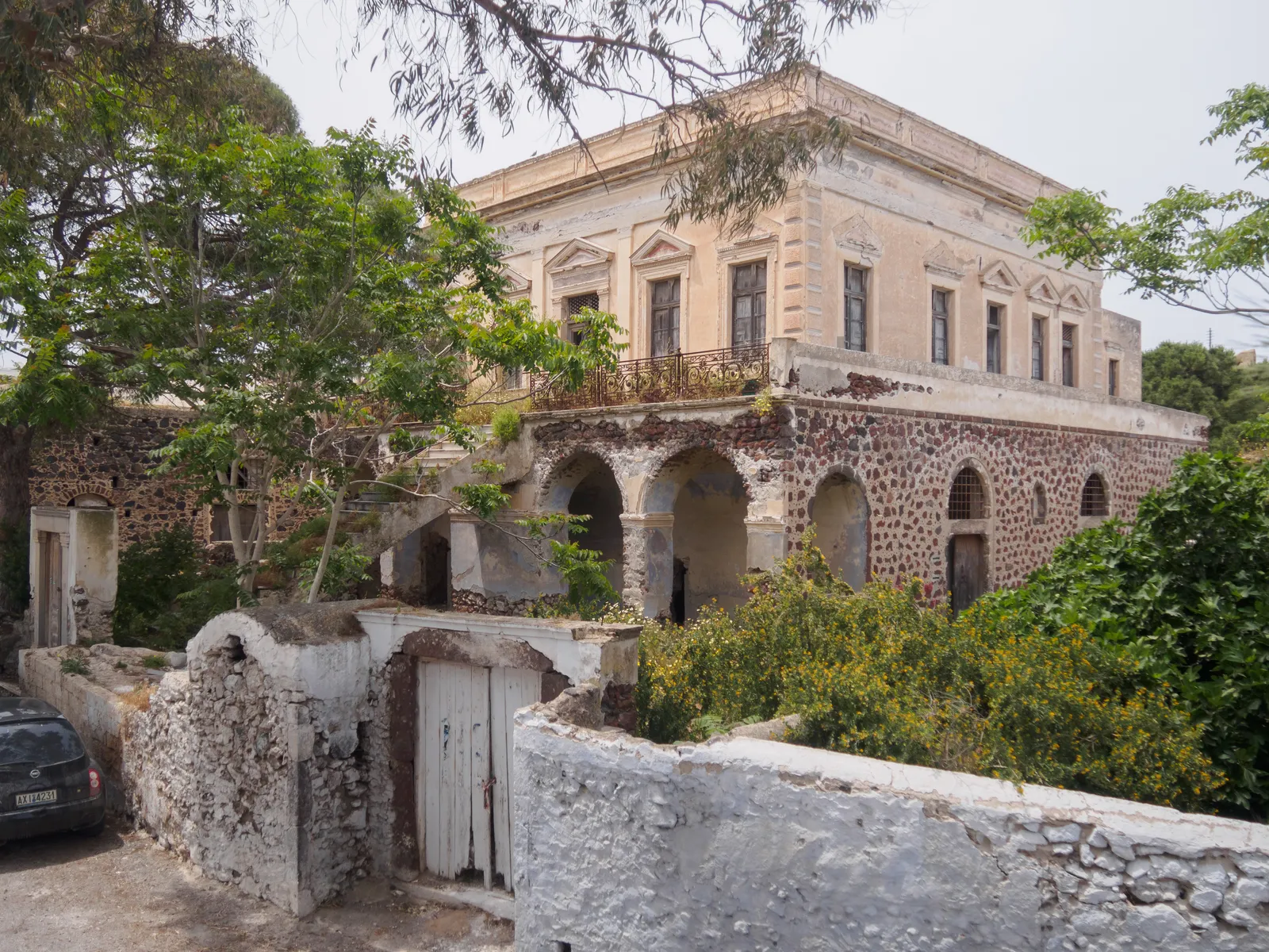 The unrestored Saliveros mansion in Messaria, a two-storey neoclassical house with peeling ochre plaster above a row of stone arches, half hidden behind overgrown trees