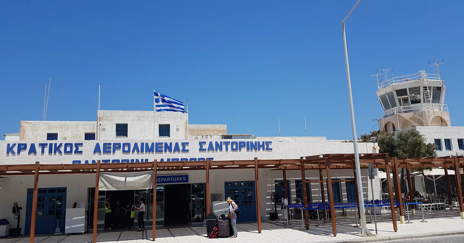 The low white frontage of Santorini's airport terminal under a Greek flag, with its name painted in Greek and English along the parapet, a wooden canopy over the departures doors and the control tower behind