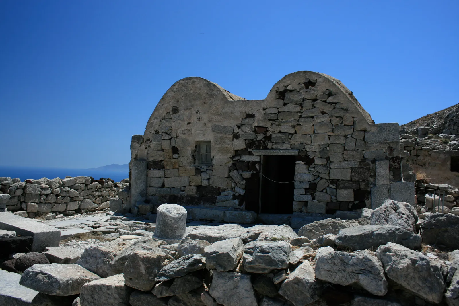 A roofless stone building at Ancient Thera with two rounded gable ends still standing, surrounded by tumbled limestone blocks and open sea on the horizon