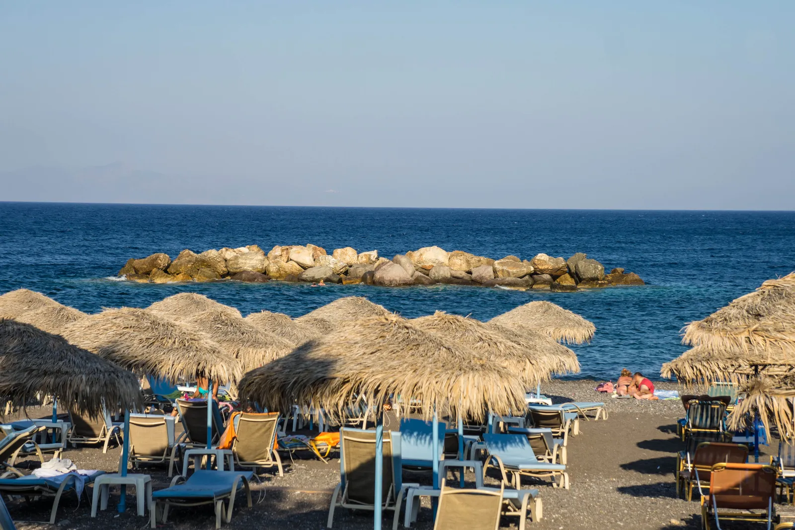 Rows of straw umbrellas and blue sunbeds on Kamari's dark grey sand, with a low rock breakwater lying just offshore in flat blue water