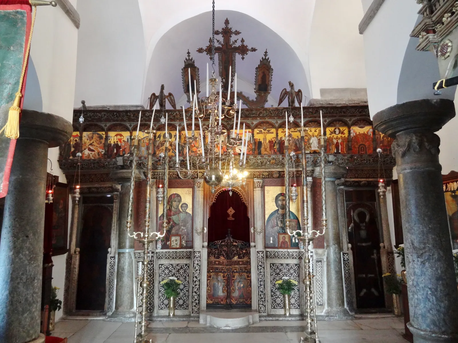 The interior of Panagia Episkopi at Mesa Gonia, a carved and gilded icon screen under a whitewashed vault with hanging brass chandeliers and dark stone columns