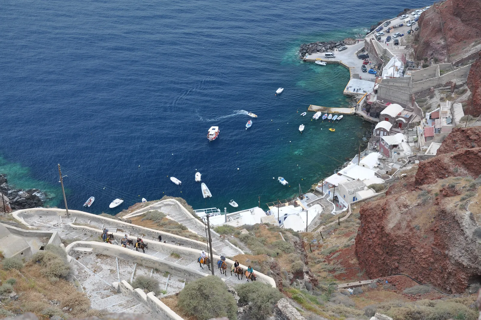Ammoudi harbour from above, small white boats moored in clear green water beside a quay of low buildings, with the zigzag stepped path and a line of donkeys descending the cliff toward it