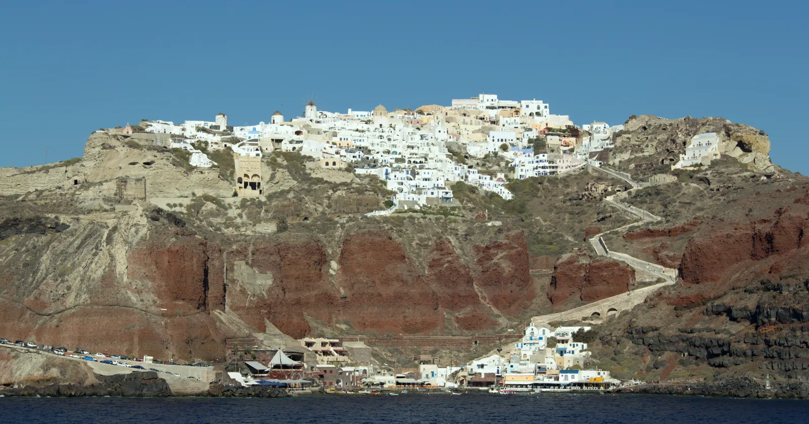Oia seen from the water, a white village along the top of a layered red and grey cliff with the small harbour of Ammoudi and its boats at the base