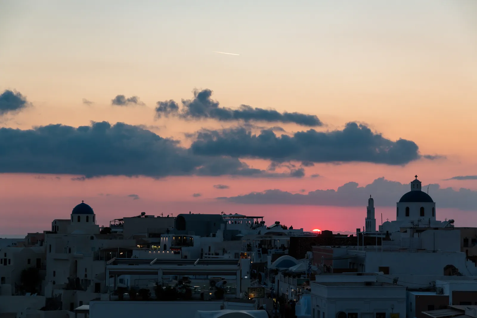 Oia at sunset, blue domes and a bell tower in silhouette against a pink and orange sky with the sun almost down behind the roofline
