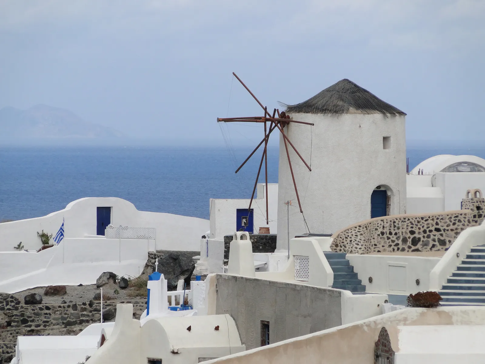 A whitewashed windmill in Oia with its bare wooden sail frame still in place and a dark conical cap, standing above the flat roofs of the village with the sea behind