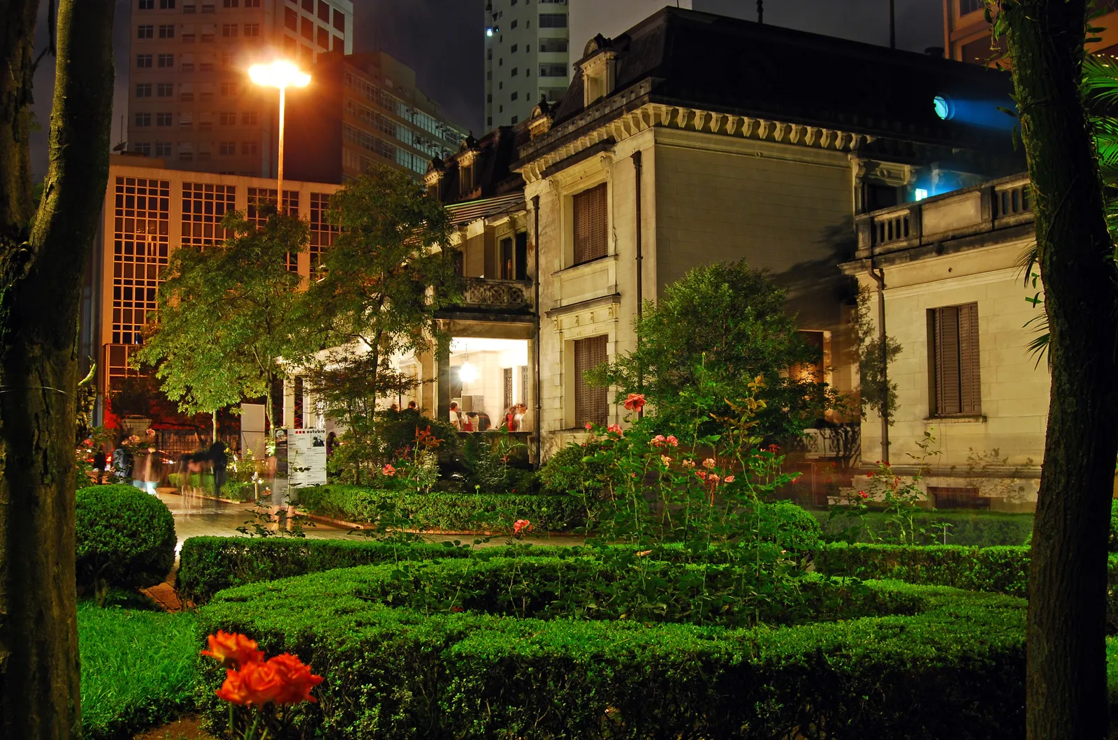 The Casa das Rosas in São Paulo lit at night, a two-storey stucco mansion behind a formal box-hedged rose garden, with the glass office towers of Avenida Paulista rising directly behind it