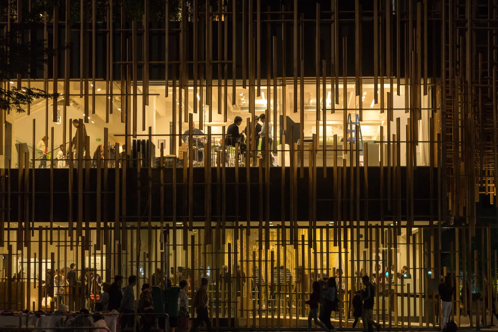 The front of Japan House in São Paulo at night, a screen of vertical timber slats across two floors of glazing with lit gallery spaces and visitors visible behind it