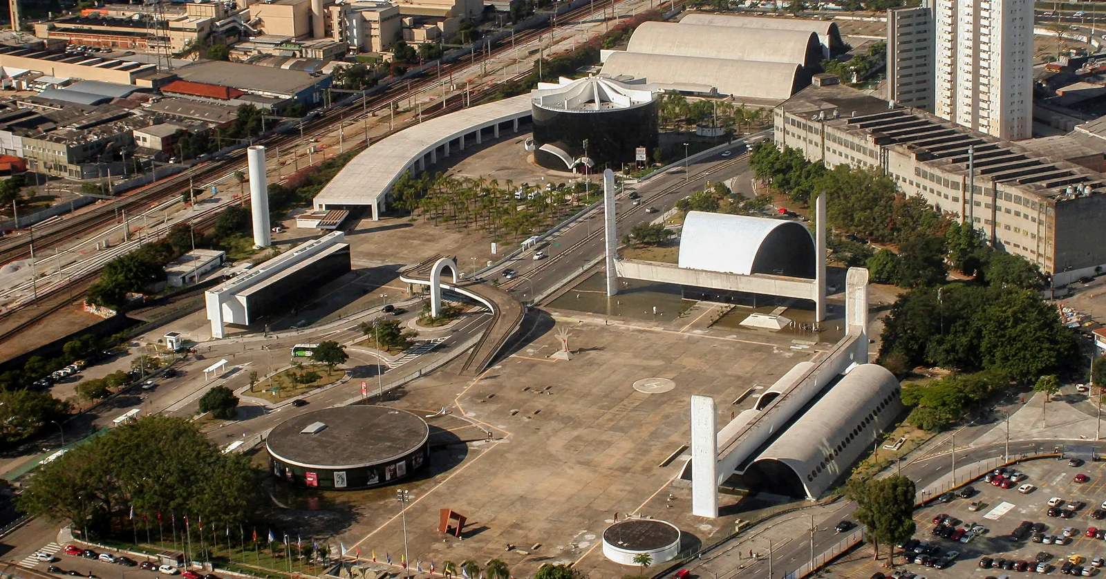The Latin America Memorial in São Paulo seen from above, Oscar Niemeyer's white curved and barrel-vaulted concrete buildings arranged around a broad empty plaza, with railway lines and city blocks behind