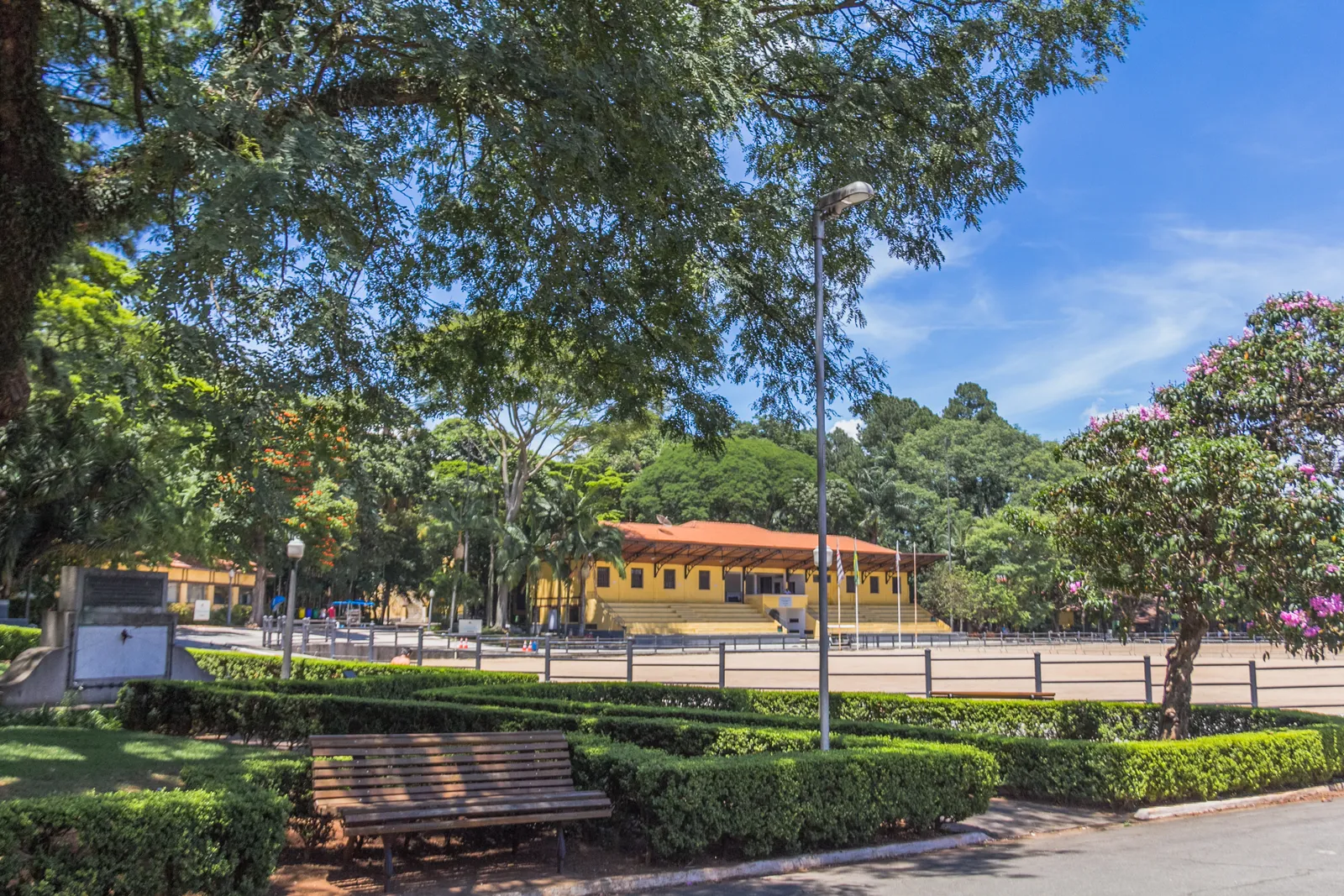 A yellow colonial-style pavilion with a red tiled roof across a sand riding arena in the Parque da Água Branca in São Paulo, with clipped hedges, benches and mature trees in the foreground