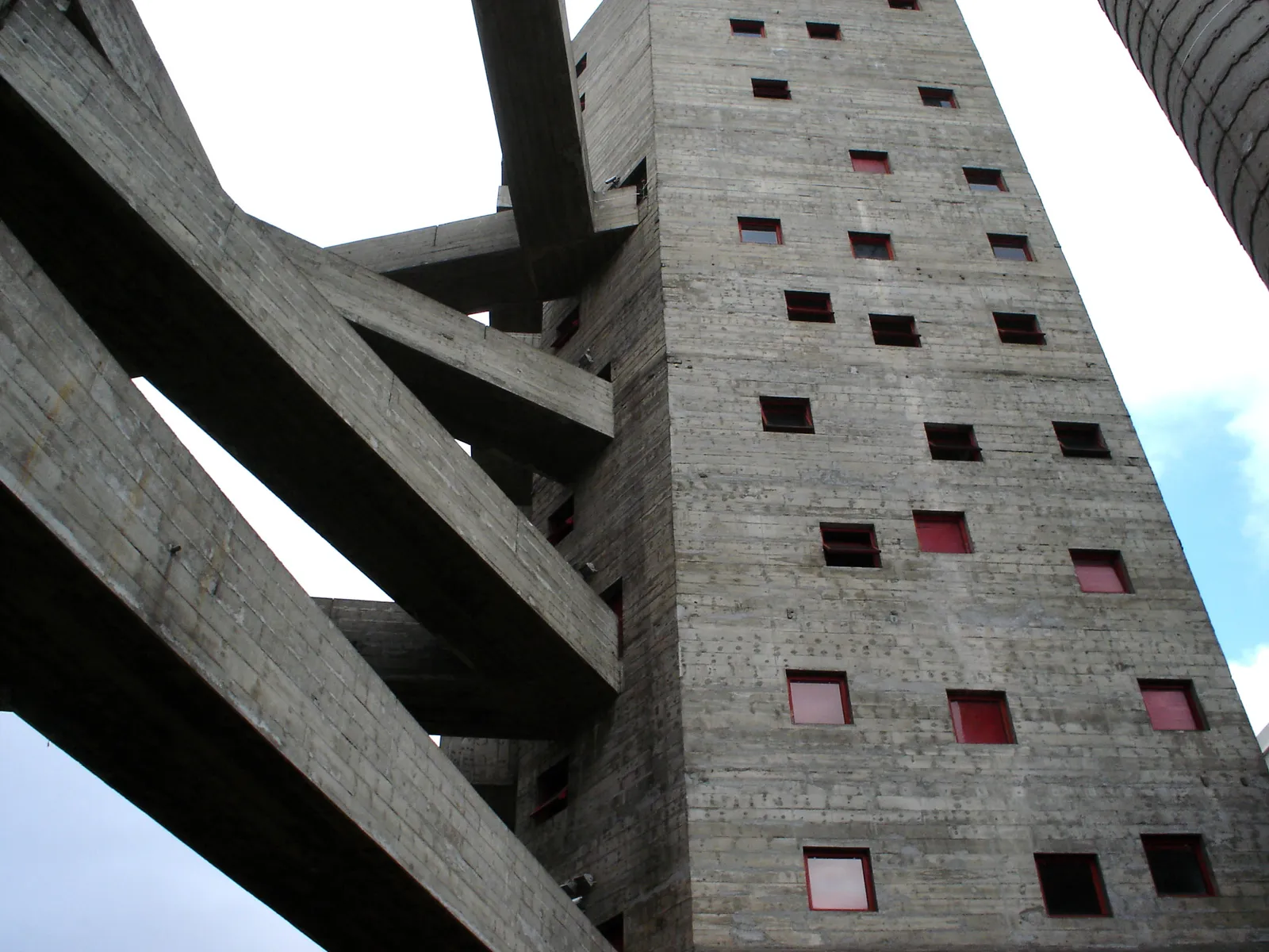 The concrete towers of Sesc Pompeia in São Paulo seen from below, raw grey walls punched with irregular red-framed openings and linked by angled walkway bridges against a pale sky