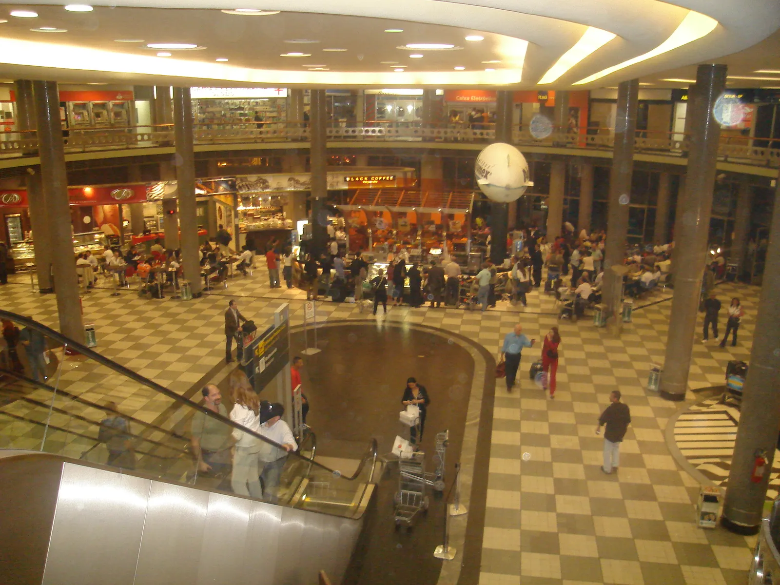The central hall of Congonhas airport in São Paulo seen from the mezzanine, with a chequered stone floor, ranks of columns, a balcony running round the upper level and passengers crossing below