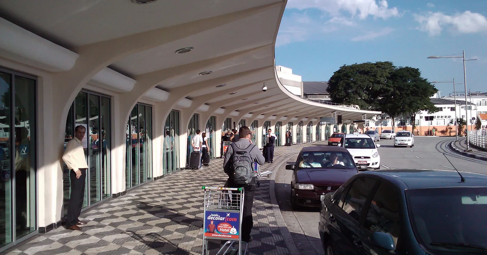 The kerbside frontage of Congonhas airport in São Paulo, a long curved white concrete canopy running above patterned Portuguese pavement, with travellers and a luggage trolley beneath it and cars parked along the road