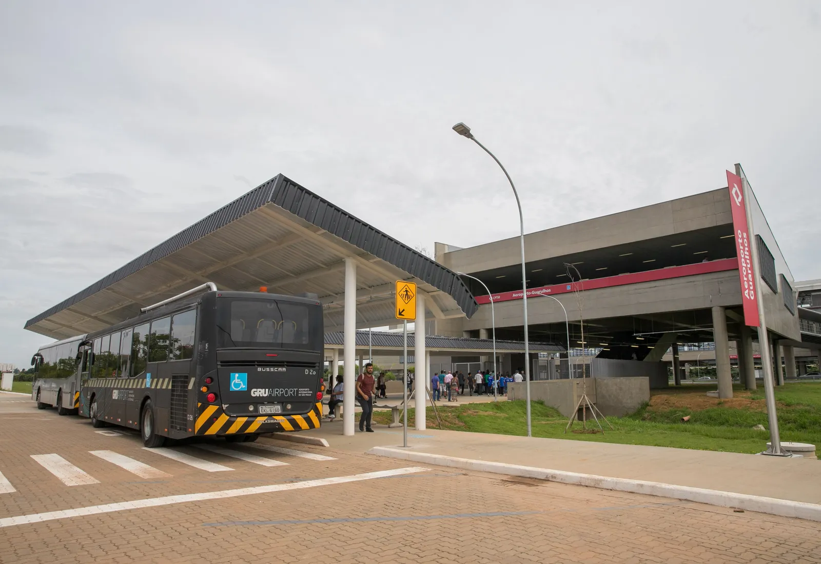 The Aeroporto-Guarulhos railway station building with a long canopied bus stand in front of it, an airport shuttle bus waiting under the canopy and passengers crossing towards the station entrance