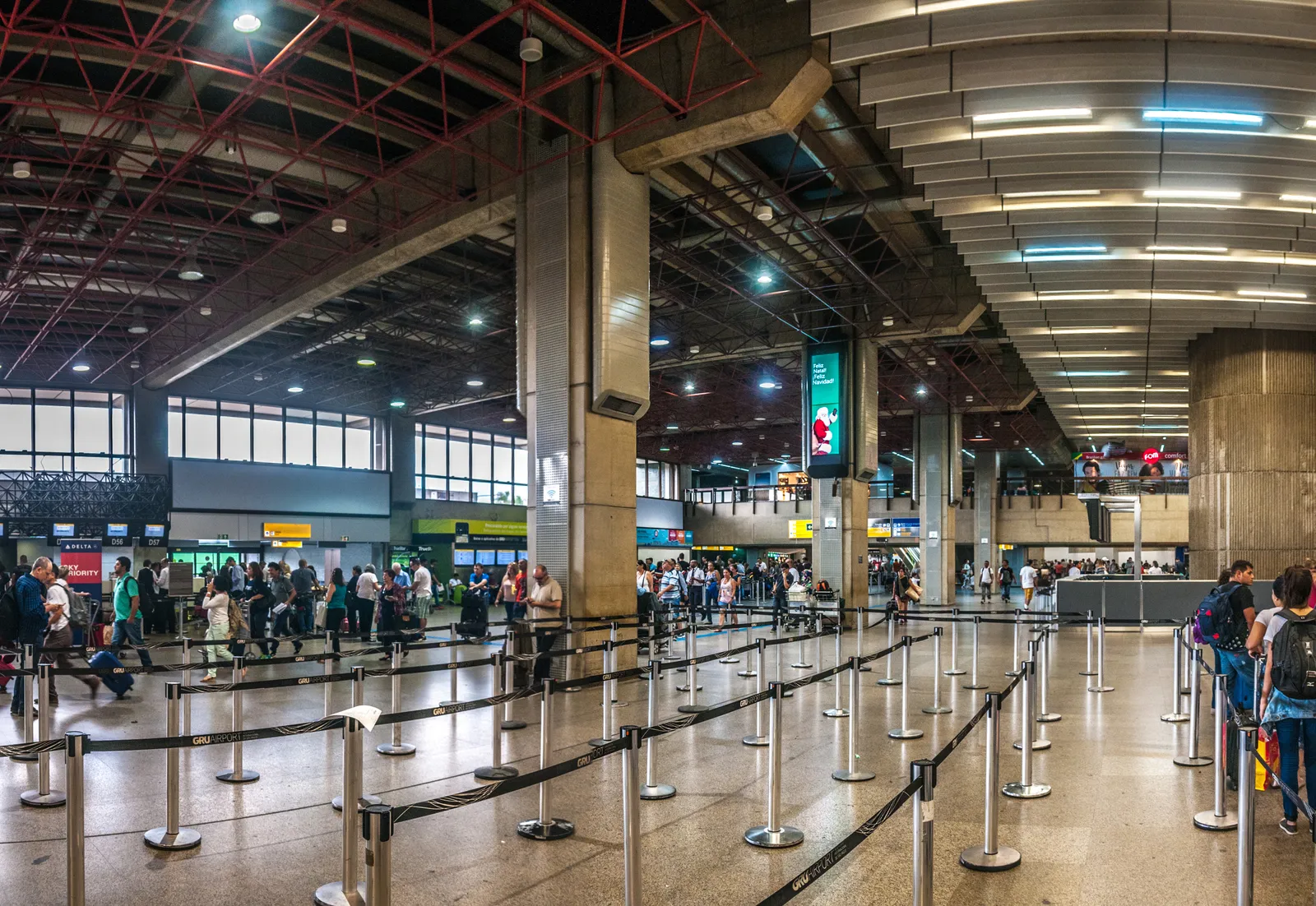 The check-in hall of Terminal 2 at Guarulhos airport in São Paulo, with heavy concrete beams overhead, ranks of empty queue barriers in the foreground and passengers lining up at the desks beyond