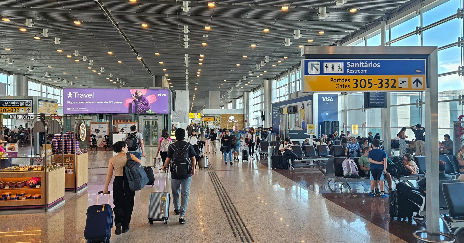 The departures concourse of Terminal 3 at São Paulo's Guarulhos airport, a wide polished-floor hall with a ribbed ceiling, gate signage for gates 305 to 332 and passengers walking past seating with wheeled suitcases