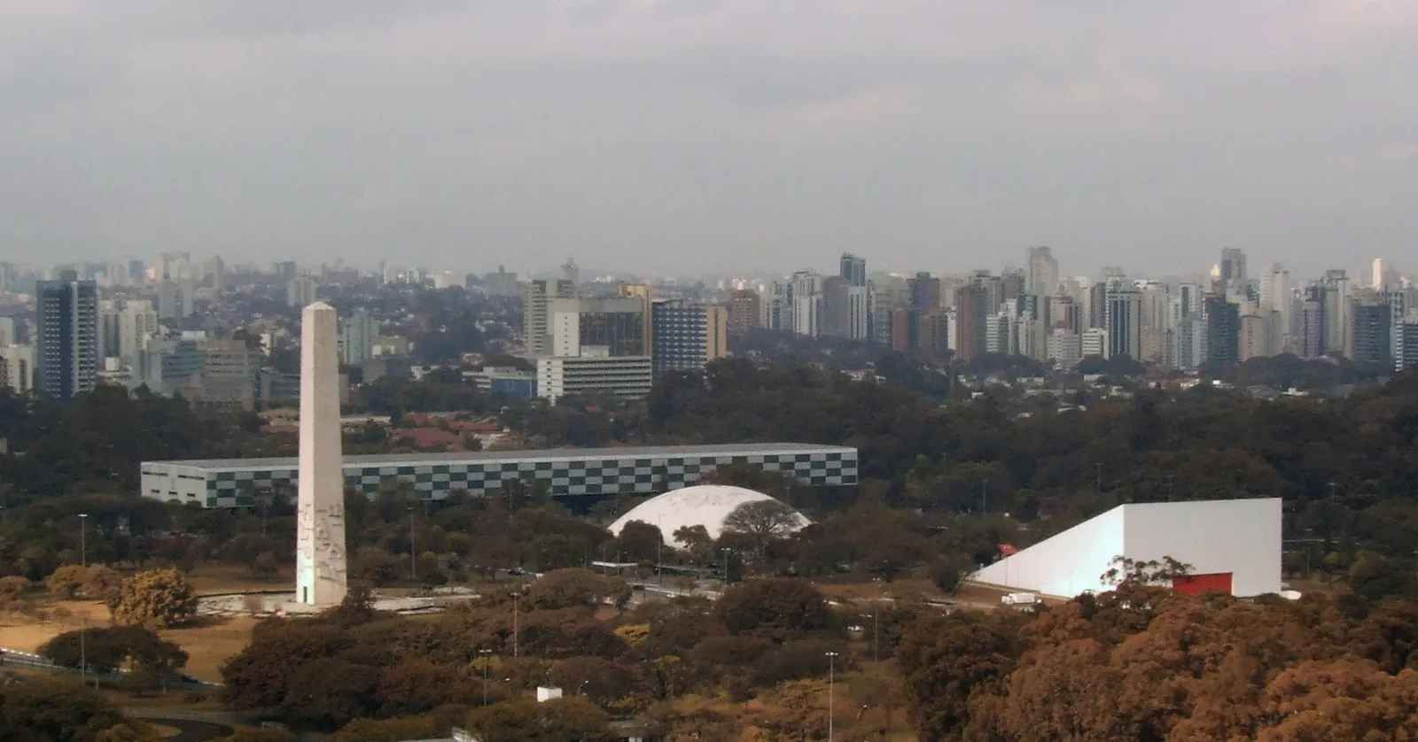 The Niemeyer buildings of Ibirapuera Park in São Paulo seen from a distance across treetops, the tall white obelisk on the left, the low chequered Bienal pavilion, the domed Oca and the white wedge of the auditorium, with the city skyline behind