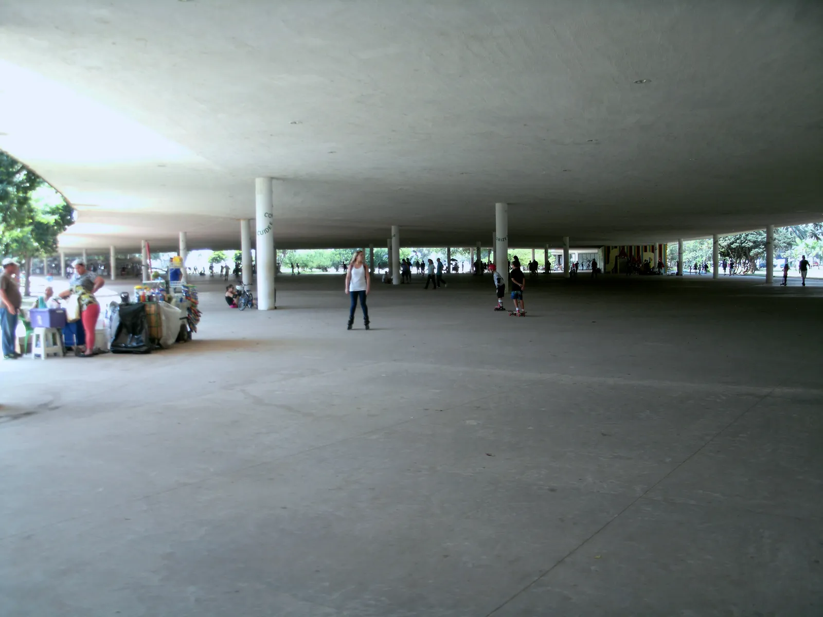 Under the concrete marquise at Ibirapuera Park in São Paulo, a wide covered deck on slender columns with stalls set up along one edge, a skater crossing the open floor and the park visible beyond the columns