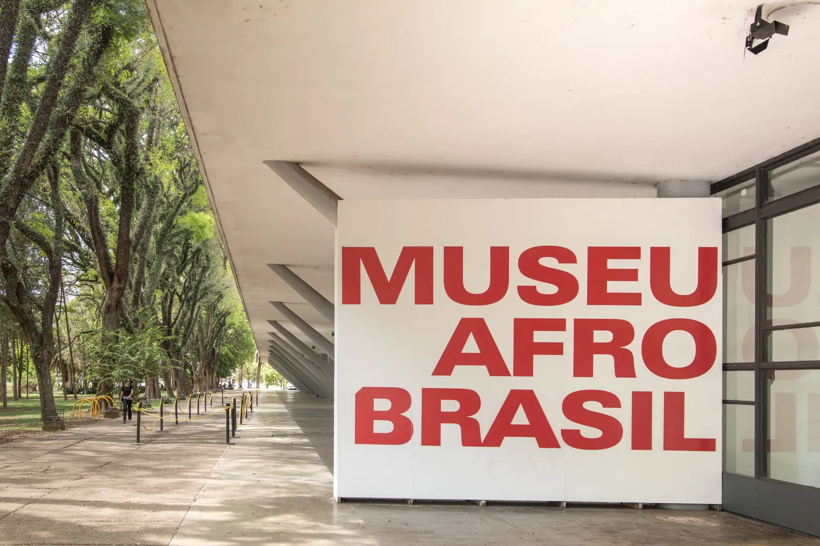 The entrance to the Museu Afro Brasil in Ibirapuera Park, a white wall carrying the museum's name in large red letters under a concrete canopy, with a tree-lined park path running alongside it