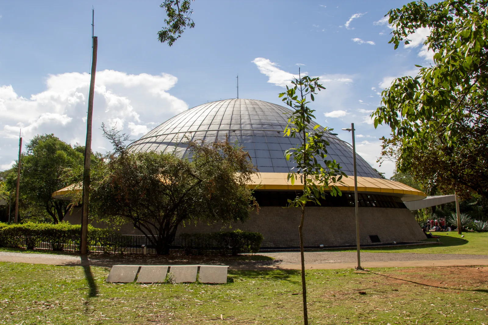 The Planetário Professor Aristóteles Orsini in Ibirapuera Park, a low circular building under a ribbed metal dome with a yellow eaves band, seen across grass and young trees