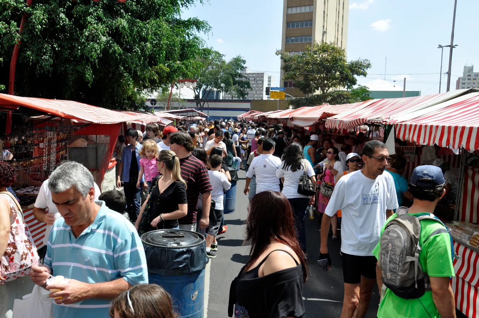 The weekend street fair at Praça da Liberdade in São Paulo, rows of red and white striped stall canopies packed tightly together with dense crowds moving between them under a bright sky