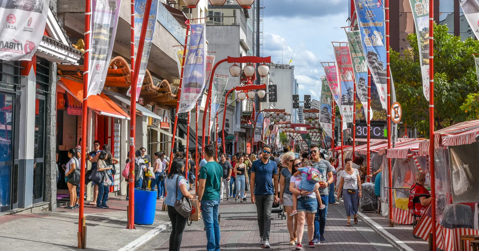 Rua Galvão Bueno in the Liberdade district of São Paulo, a pedestrianised street lined with tall red lamp posts and hanging banners, market awnings along both sides and crowds walking down the middle