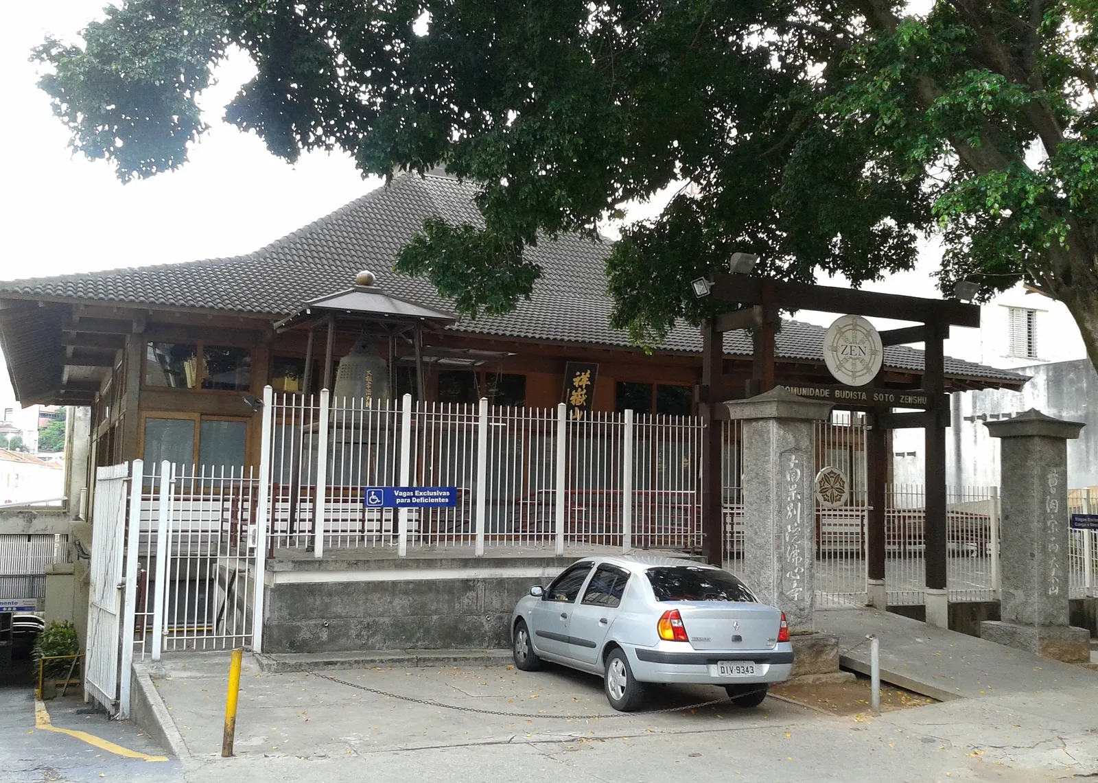 The Busshinji temple on Rua São Joaquim in São Paulo, a low timber building with a broad pyramidal tiled roof and a hanging bell under the eaves, behind railings and a stone gate post