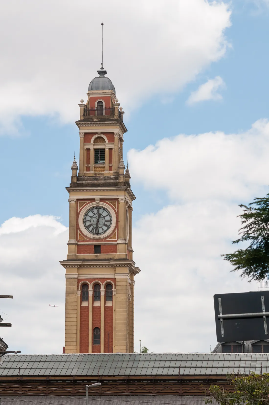 The clock tower of Luz Station in São Paulo rising above the station roof against a blue sky, its brick and stone stages topped by a small dome, with a white clock face on the front