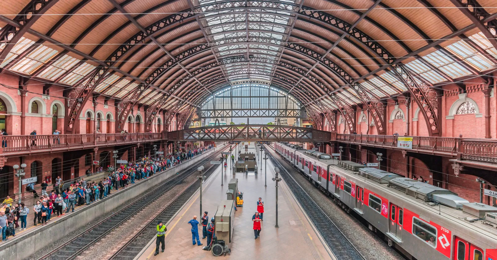 The interior of Luz Station in São Paulo, a wide platform hall under an arched iron and glass roof with pink brick walls on both sides, a red commuter train at one platform and passengers waiting along another