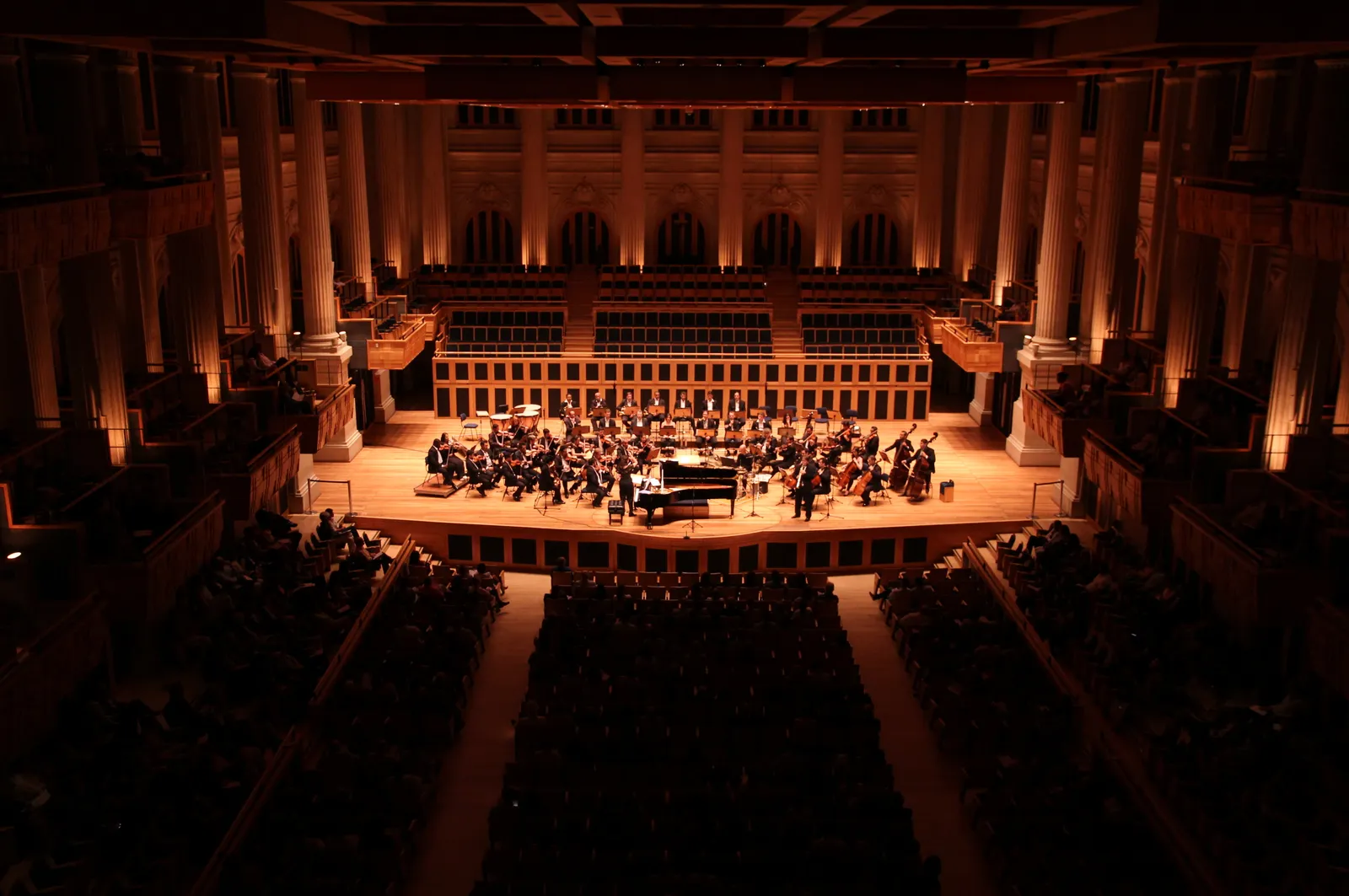 The main hall of Sala São Paulo seen from a rear balcony during a concert, an orchestra on the platform under warm light with tall columns and coffered walls rising around a full audience