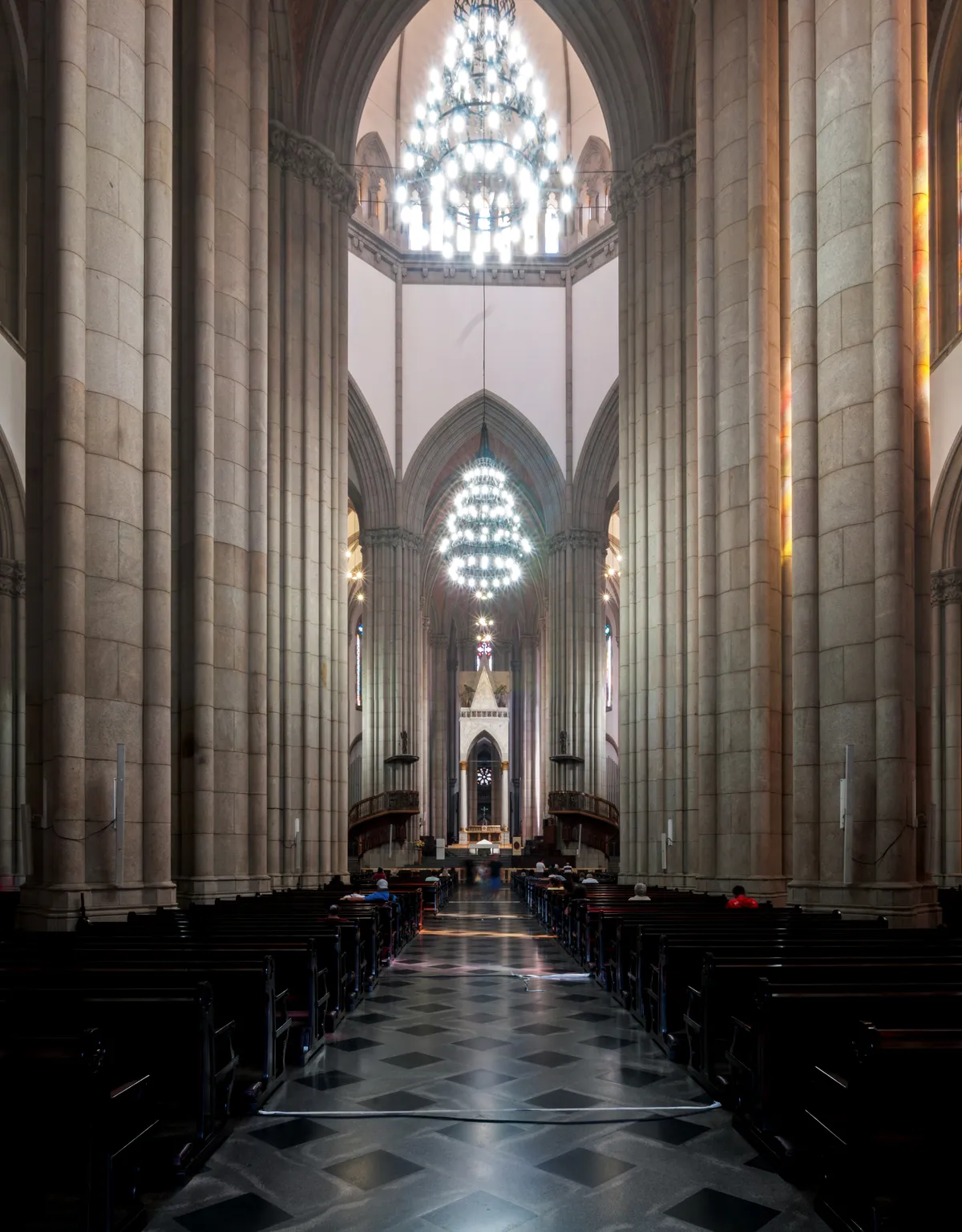 The nave of the Sé cathedral in São Paulo, tall pale stone columns rising to pointed arches with chandeliers hanging between them and a black-and-white chequered marble floor running towards the altar