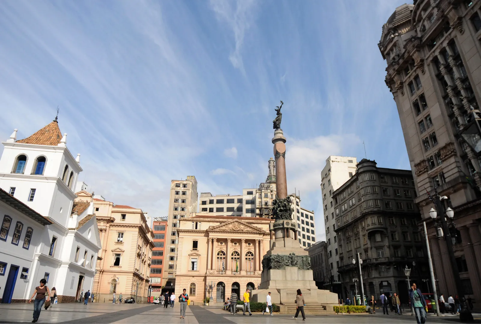 The open square at Pátio do Colégio in São Paulo, with the whitewashed Jesuit church and its tower on the left, a tall stone monument column in the centre and ornate early 20th-century buildings around the edges
