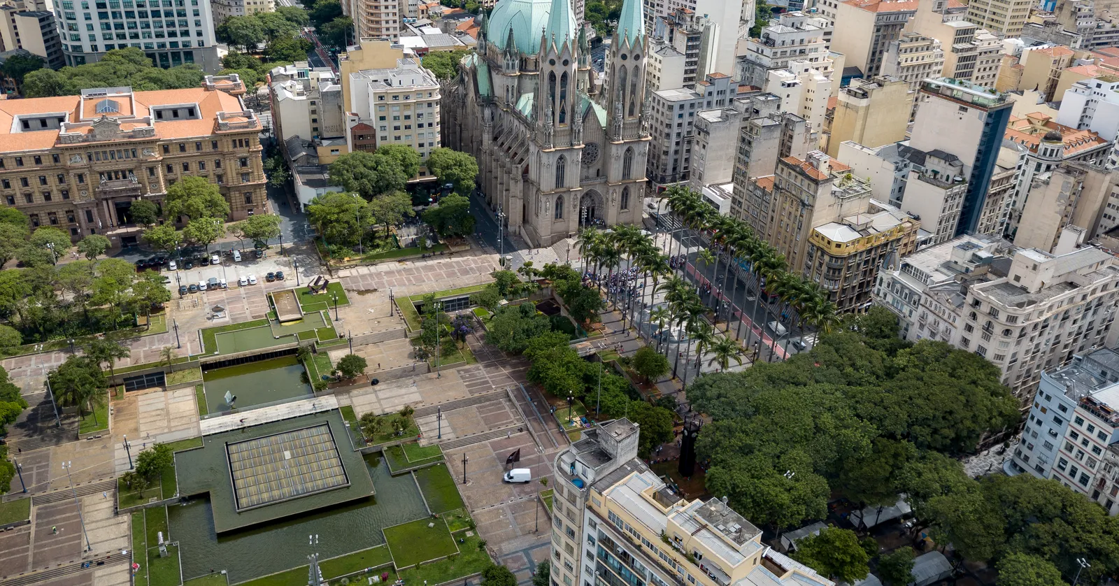 Praça da Sé in São Paulo seen from above, the twin-towered cathedral standing on one side of a long paved square lined with palms and reflecting pools, surrounded by dense city blocks