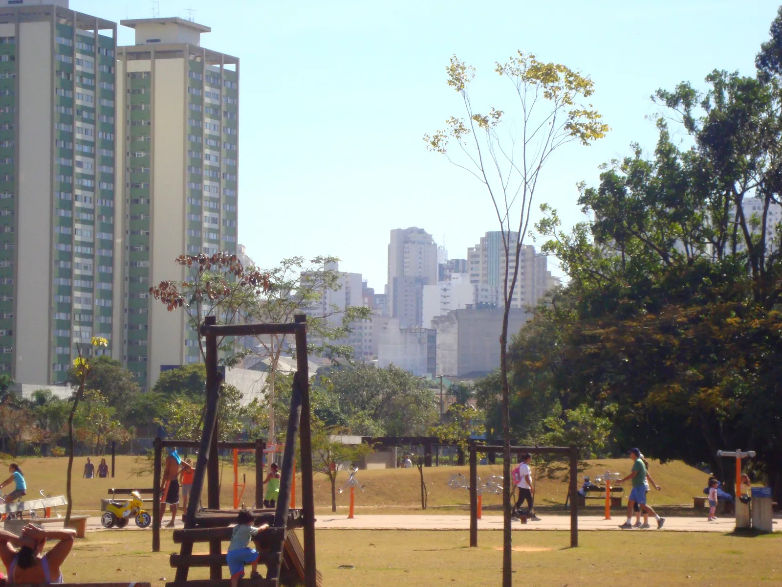 Open lawns and play equipment in the Parque da Juventude in northern São Paulo, with people walking and exercising on the grass and tall apartment towers rising along the far edge of the park