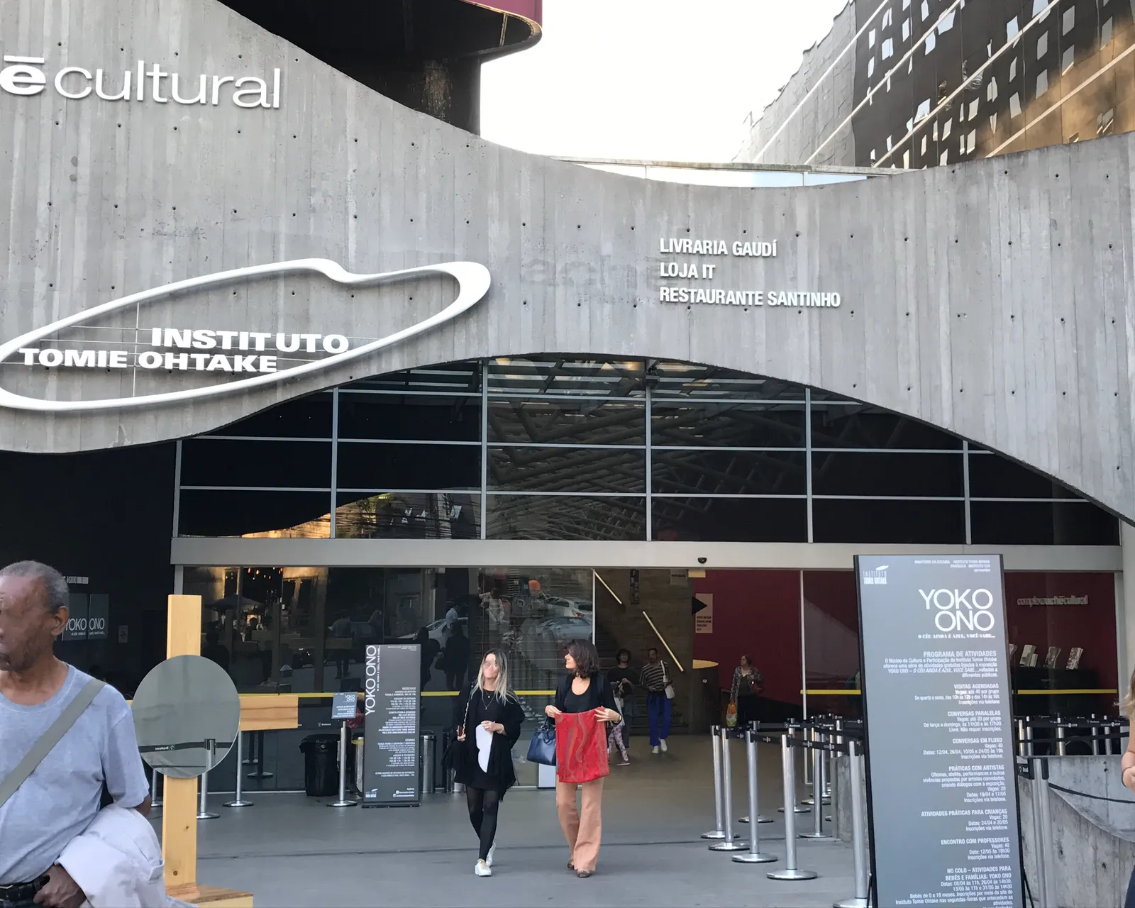 The entrance to the Instituto Tomie Ohtake in São Paulo, a curved concrete canopy carrying the institute's name in white lettering above glass doors, with visitors walking out onto the forecourt