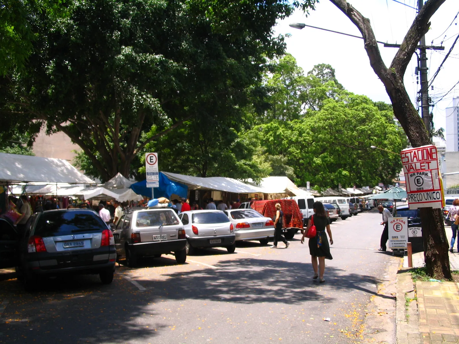 Stalls with white canopies set up under mature trees along the edge of Praça Benedito Calixto in São Paulo, with parked cars, browsing shoppers and a leafy residential street beyond