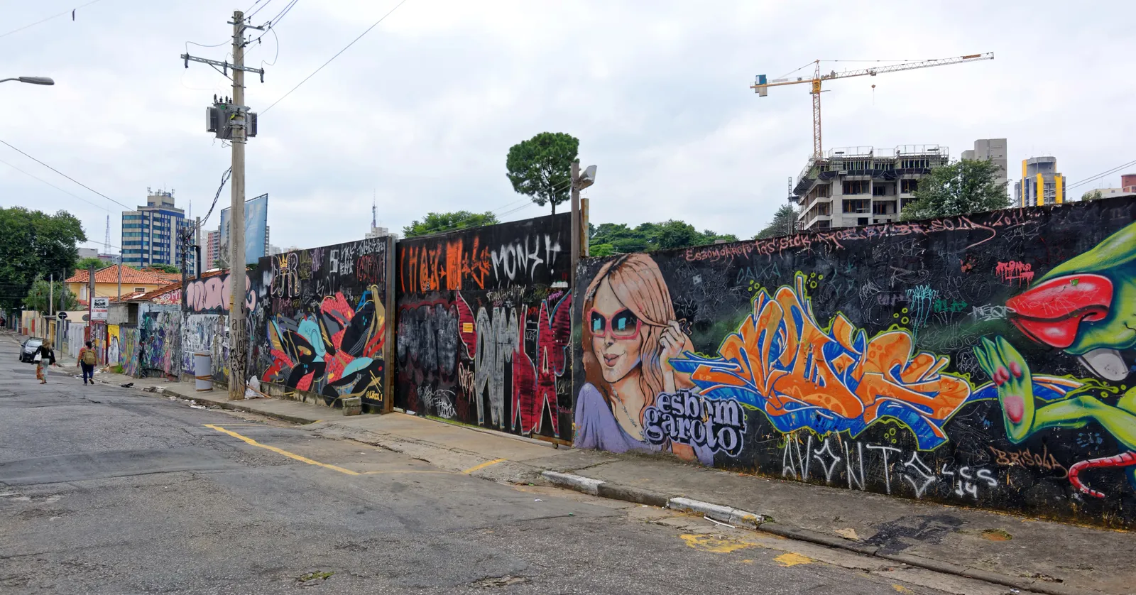 A side street in Vila Madalena, São Paulo, with a long wall painted end to end in murals running down one side, the empty roadway and kerb in front of it and apartment blocks and a construction crane on the skyline