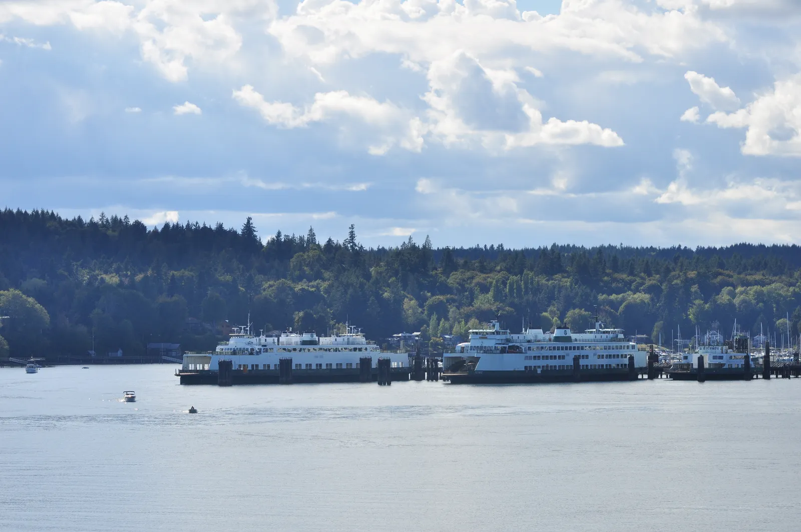 Two white and green Washington State ferries tied up at the wooded Bainbridge Island terminal across the water, under a blue sky with scattered cloud