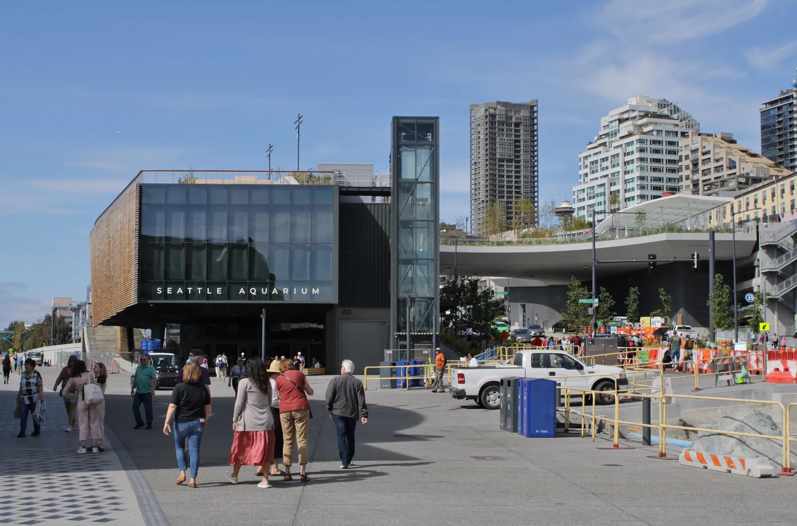 The glass and timber front of the Seattle Aquarium's Ocean Pavilion on the downtown waterfront, with people walking past on the wide promenade and apartment towers behind