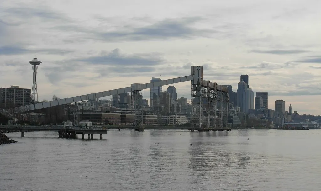 The long conveyor of the Pier 86 grain terminal reaching out over Elliott Bay from Myrtle Edwards Park in Seattle, with the Space Needle and the downtown towers behind under a grey sky